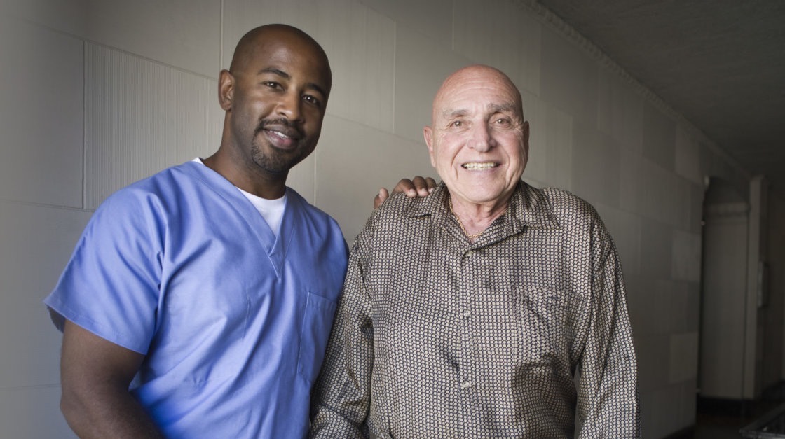 A male caregiver in scrubs with his arm around a smiling senior man