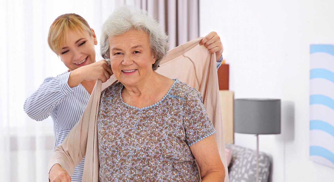 A caregiver helping a smiling senior woman put on her cardigan at home