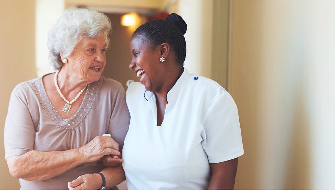 A caregiver and a smiling senior woman walking arm in arm at home