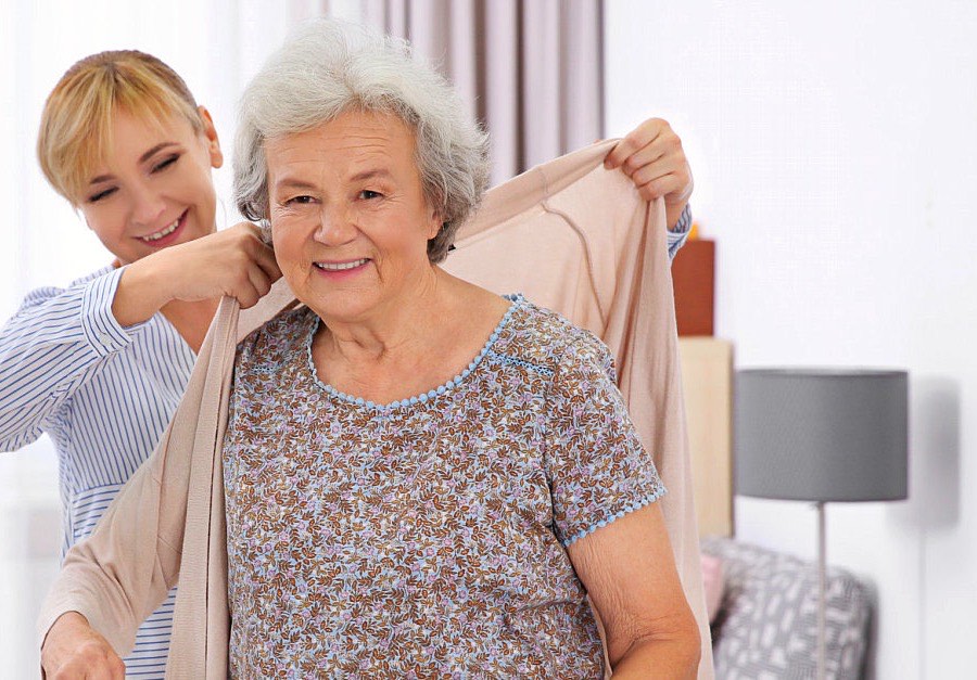 A caregiver helping a smiling senior woman put on her cardigan at home